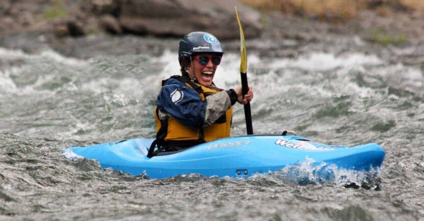 A person in a blue kayak wearing a helmet and life jacket paddling through whitewater rapids, smiling energetically